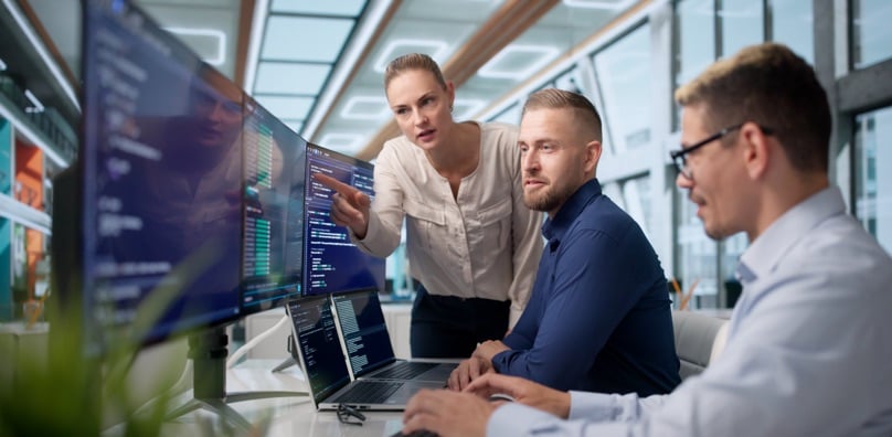 Tre colleghi analizzano righe di codice su vari monitor per gestire la sicurezza e il backup dati aziendali.