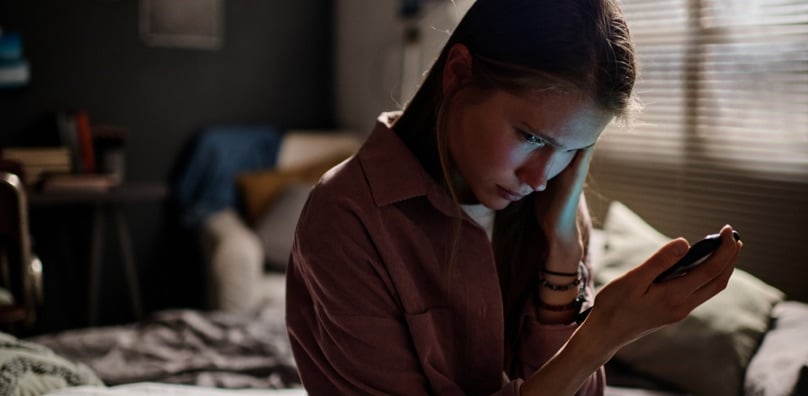 Ragazza triste in camera guarda lo smartphone, vittima di cyberbullismo, un fenomeno da cui tutelare i figli.