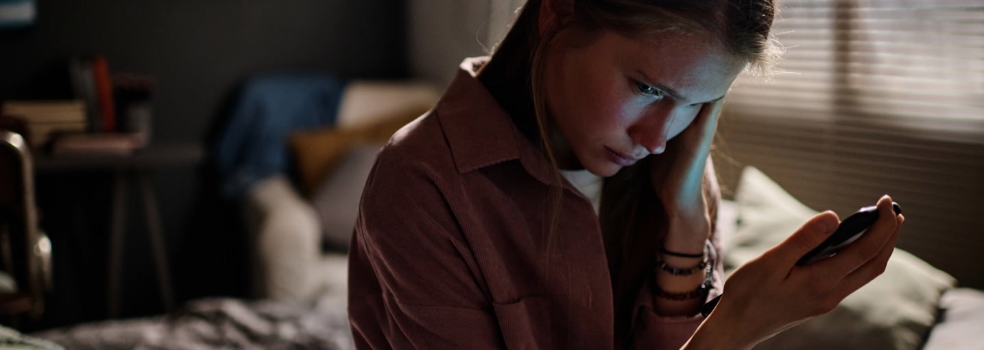 Ragazza triste in camera guarda lo smartphone, vittima di cyberbullismo, un fenomeno da cui tutelare i figli.
