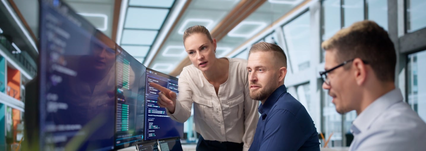 Tre colleghi analizzano righe di codice su vari monitor per gestire la sicurezza e il backup dati aziendali.