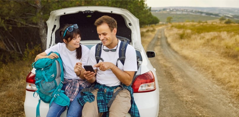 Giovani viaggiatori con zaini che guardano il telefono per risparmiare strada e tappe durante le vacanze in auto.